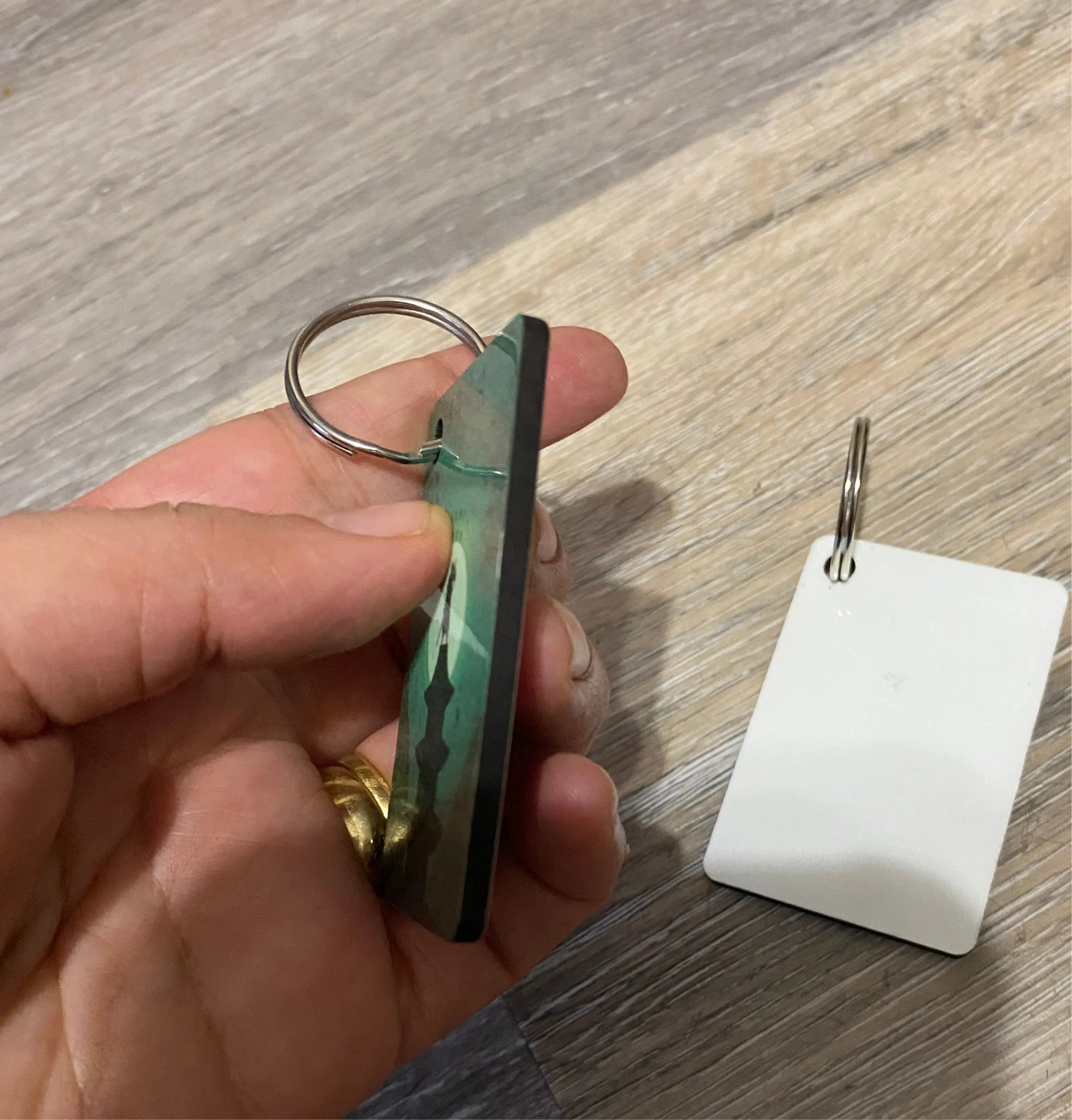 Hand holding a camouflage-patterned keychain on a wooden surface with another keychain in the background.